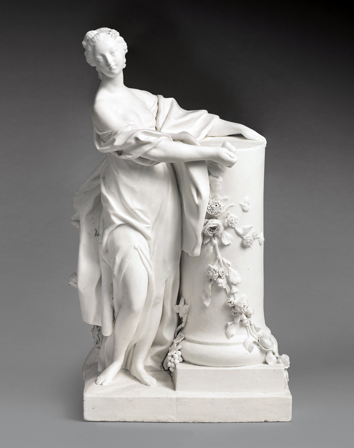 A white marble statue depicts a woman draped in flowing garments, one breast exposed, leaning slightly against a cylindrical pedestal adorned with floral carvings. The woman's hair is styled back, and her expression appears serene and contemplative.