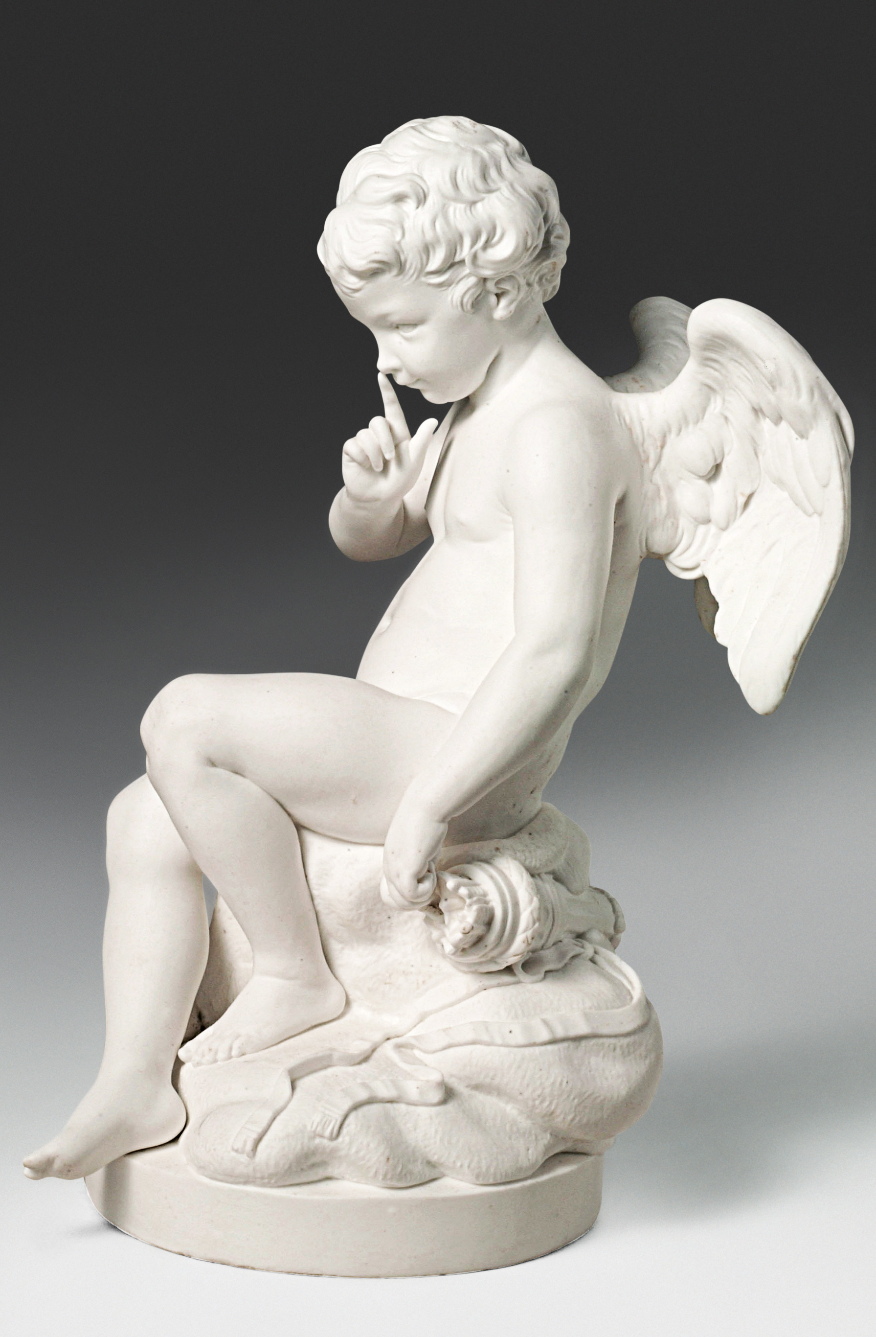 A white marble statue depicts a young cherub with wings, seated on a rock. The cherub has one finger raised to its lips, suggesting a shushing gesture. The intricately detailed figure showcases classical artistry in its form and features.