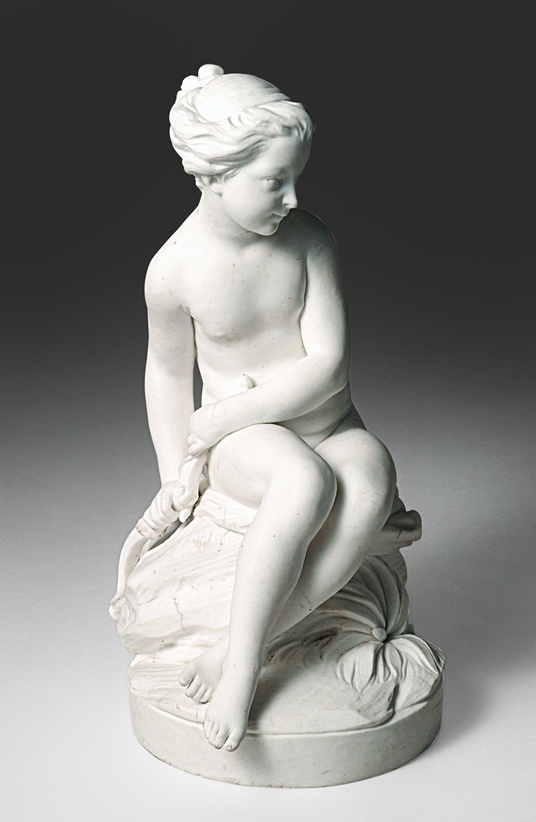 A white marble statue depicts a seated woman with her upper body modestly covered by one arm. The figure has her head turned to the side and legs crossed, with one hand resting on her lap and the other holding a piece of fabric. The base has a draped cloth detail.