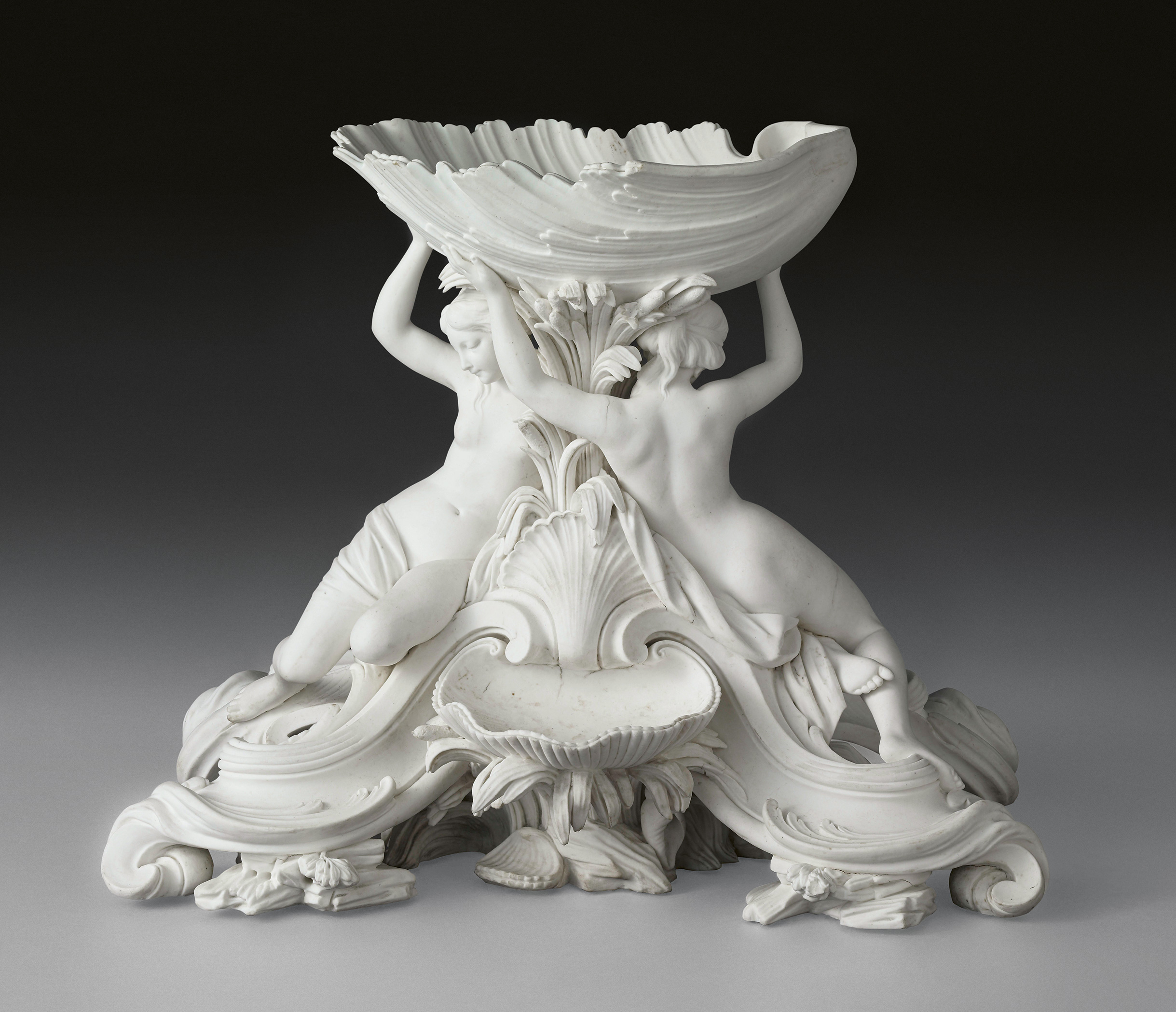 A detailed porcelain sculpture depicts two female figures back-to-back, supporting a large, shell-like bowl above them. The figures are surrounded by intricate leaf motifs and seashell designs, giving the piece an ornate and baroque appearance.