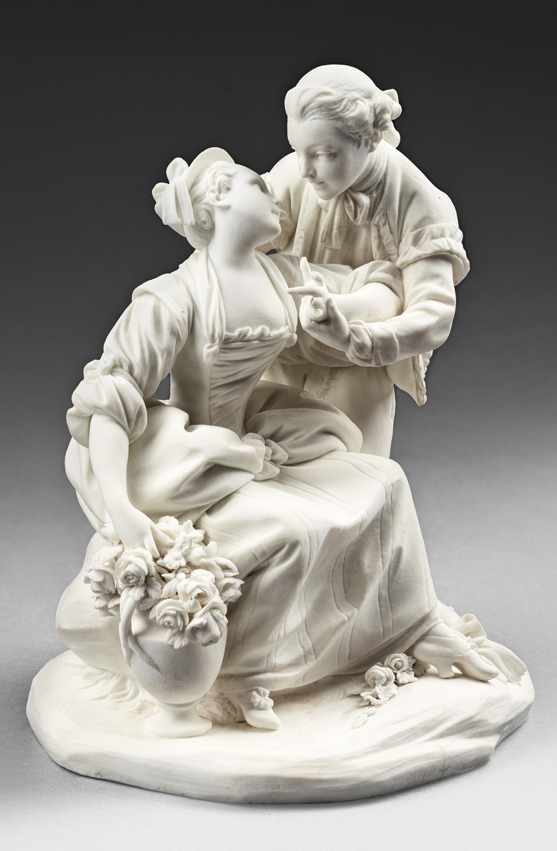 A finely crafted porcelain sculpture depicts a romantic scene. A man gently embraces a woman as she leans towards him, holding a flower bouquet. The detailed attire suggests a historical period, with attention to the draping fabric and expressive figures.