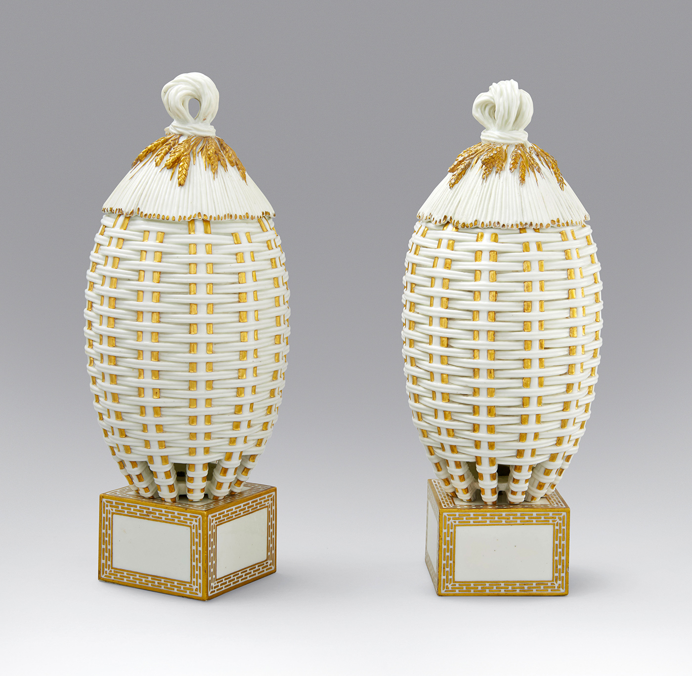 Two intricately designed porcelain baskets are displayed, each featuring a woven pattern with gold and white detailing. The baskets have rounded tops adorned with looped knots and rest on square bases, also decorated with gold accents. The background is gray.