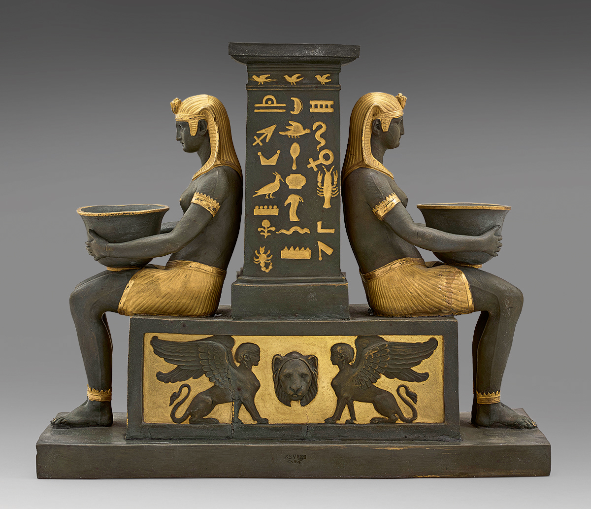 A decorative piece featuring two identical seated Egyptian-style figures with headdresses and skirts, holding bowls, positioned back-to-back. They are flanked by a central column adorned with hieroglyphics and symbols. The base has winged sphinxes and a lion face.