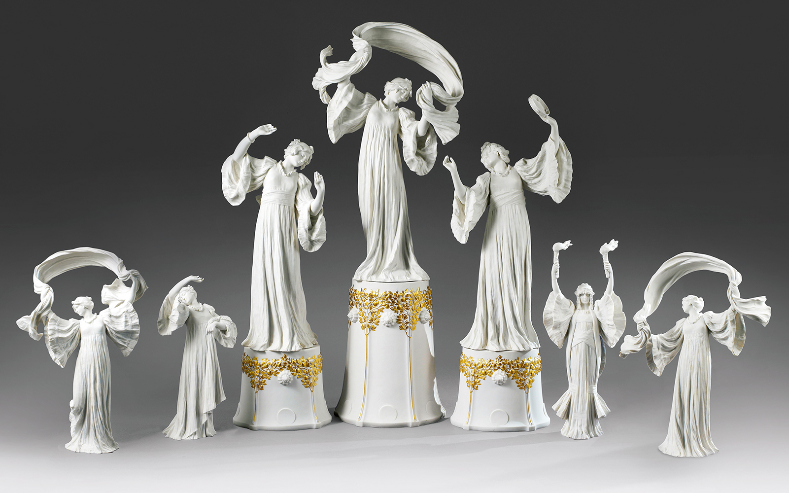 A set of intricately designed white porcelain figurines depicting elegant, flowing dancers. The central figure is elevated on a pedestal adorned with golden accents, surrounded by smaller figures in various graceful poses, each holding delicate, ribbon-like accessories.