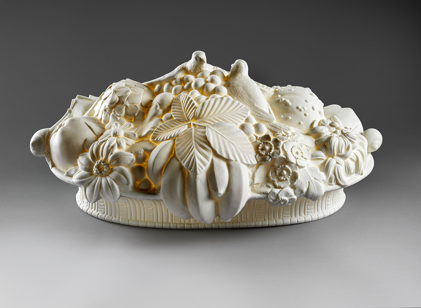 A detailed white ceramic sculpture depicting an assortment of fruits, flowers, and leaves arranged in a domed shape. The intricate design includes various textures and patterns, creating a visually rich and ornate piece.