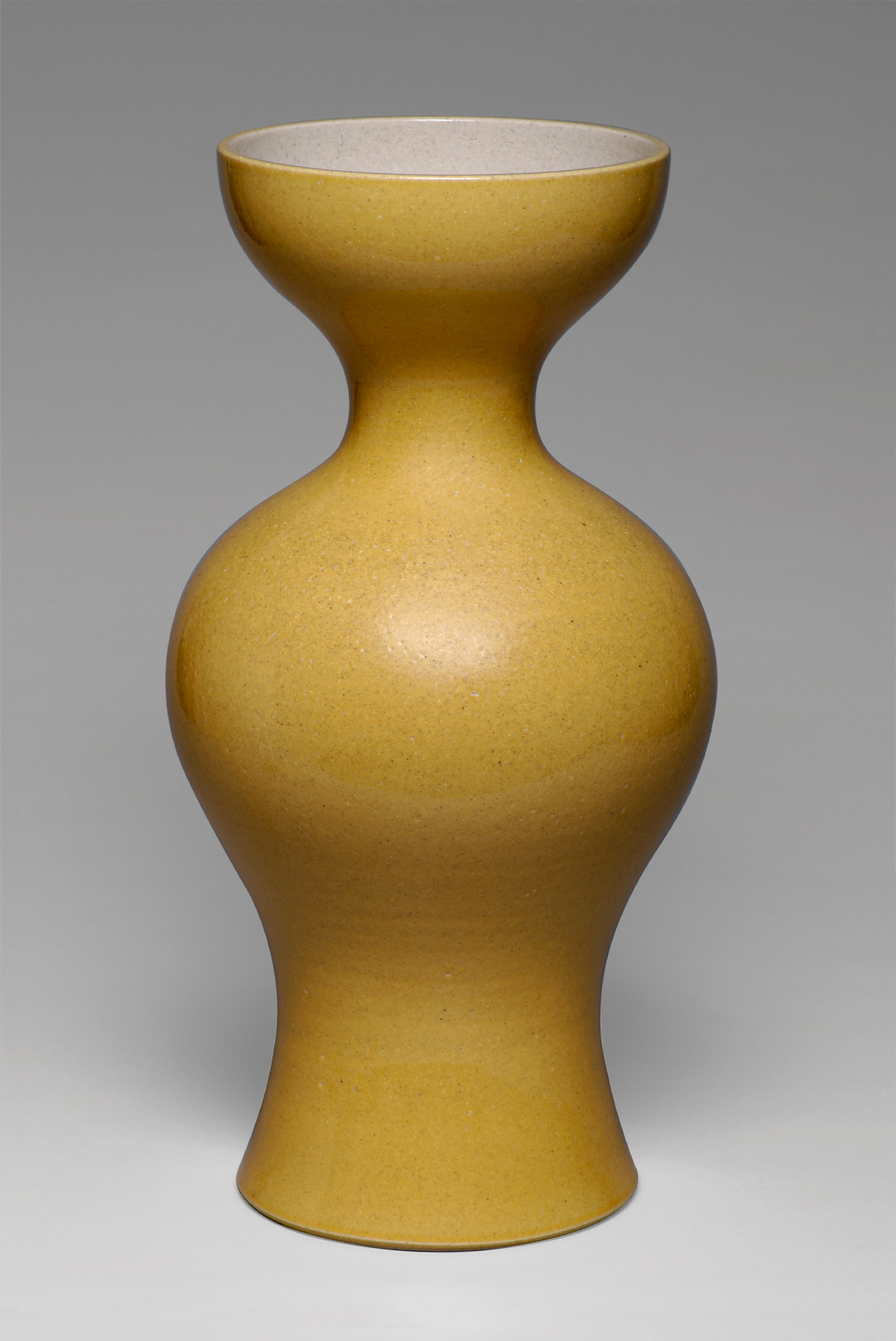 A modern, mustard-yellow, ceramic vase with a smooth, matte finish. The vase has an hourglass shape, wide at the top and bottom with a narrowed middle section. The background is a plain, light gray.