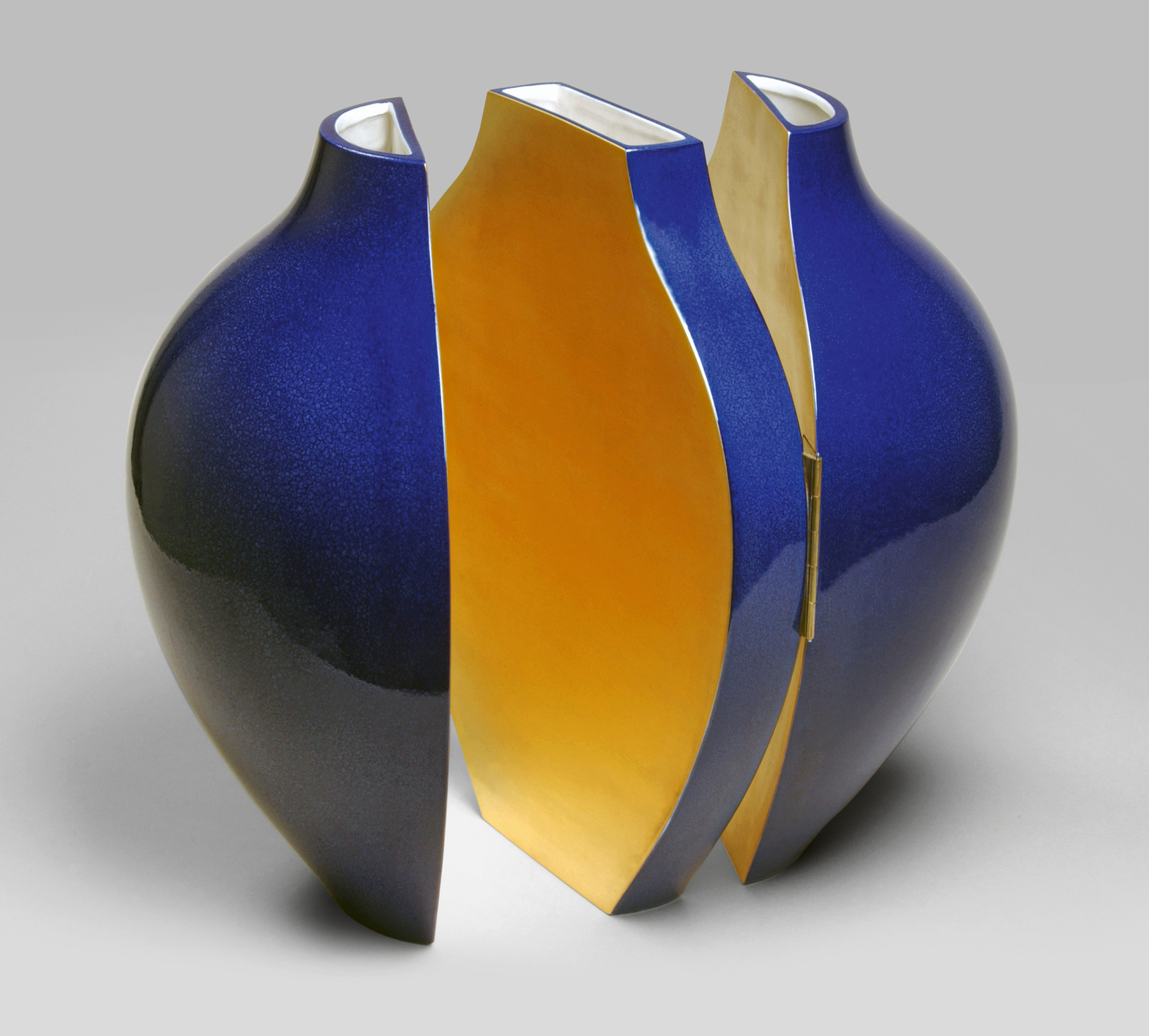 A contemporary art sculpture featuring a geometric design with two outer pieces in glossy blue and a central piece in vibrant gold. The central piece is partially concealed by the blue parts, creating a dynamic, segmented form. The sculpture appears on a neutral background.