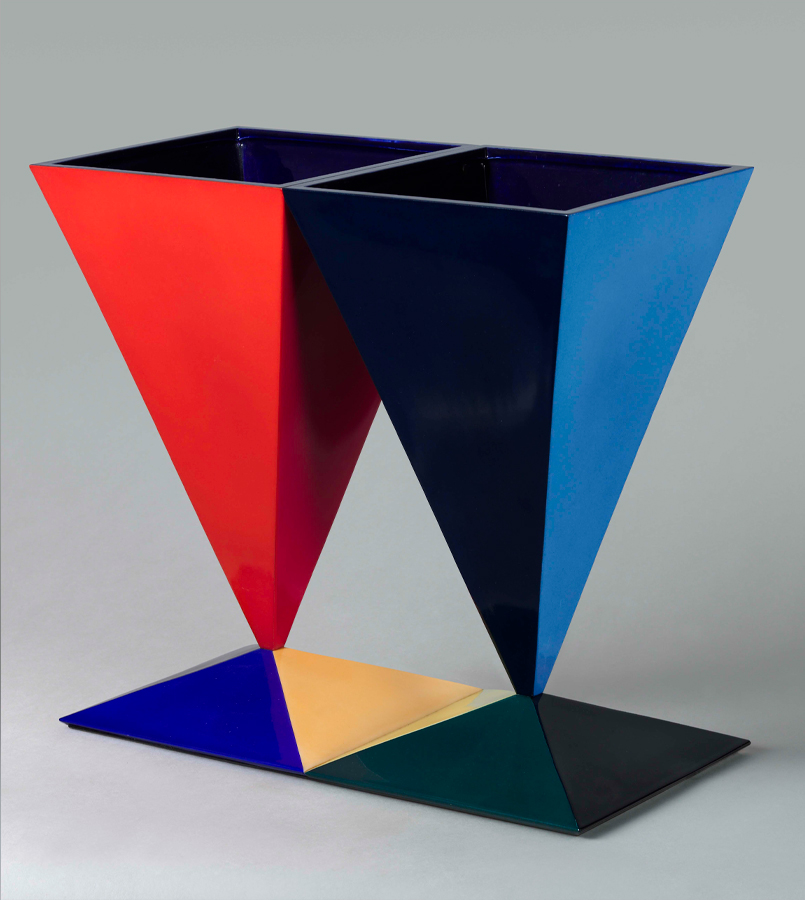 Two inverted pyramid-shaped containers, one red and one blue, are placed side by side on an abstract base. The base is divided into four colored sections: red, blue, yellow, and green. The background is plain light gray.