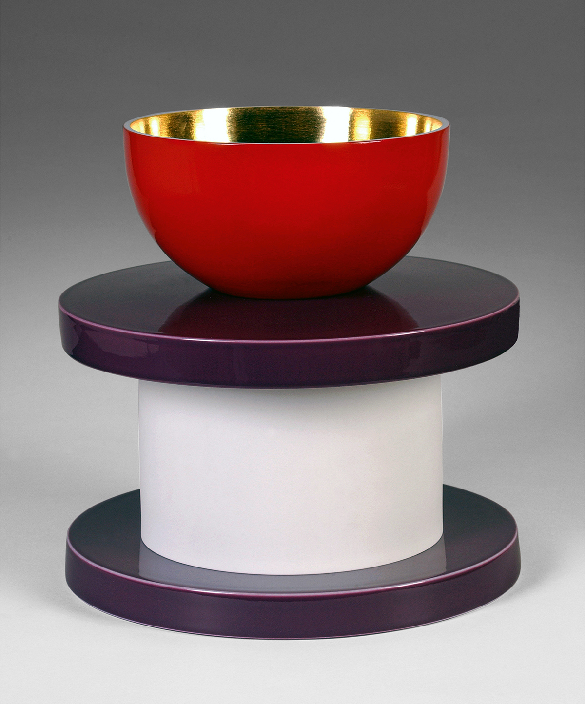 A visually striking, two-tiered stand holds a vibrant red and gold bowl. The stand has a cylindrical white center, sandwiched between two deep purple, round platforms. The bowl's shiny red exterior contrasts with its metallic gold interior, enhancing the modern aesthetic.