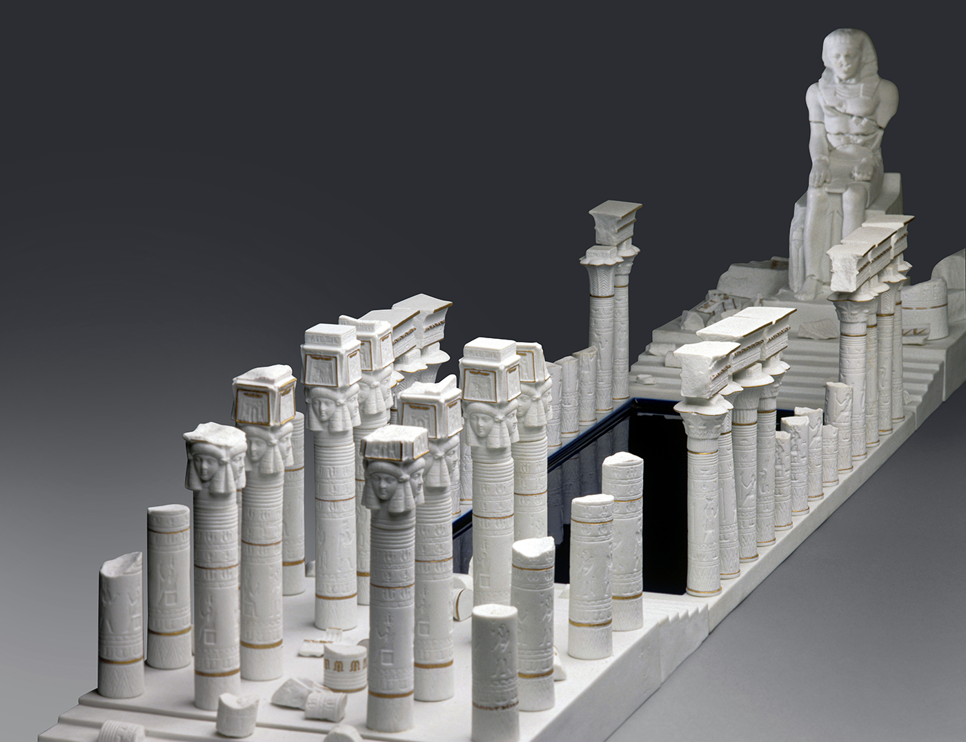 A detailed white architectural model of an ancient temple complex, featuring rows of intricately carved columns with ornate capitals and a large sculpted figure sitting at the end. The model is set against a neutral gray background.