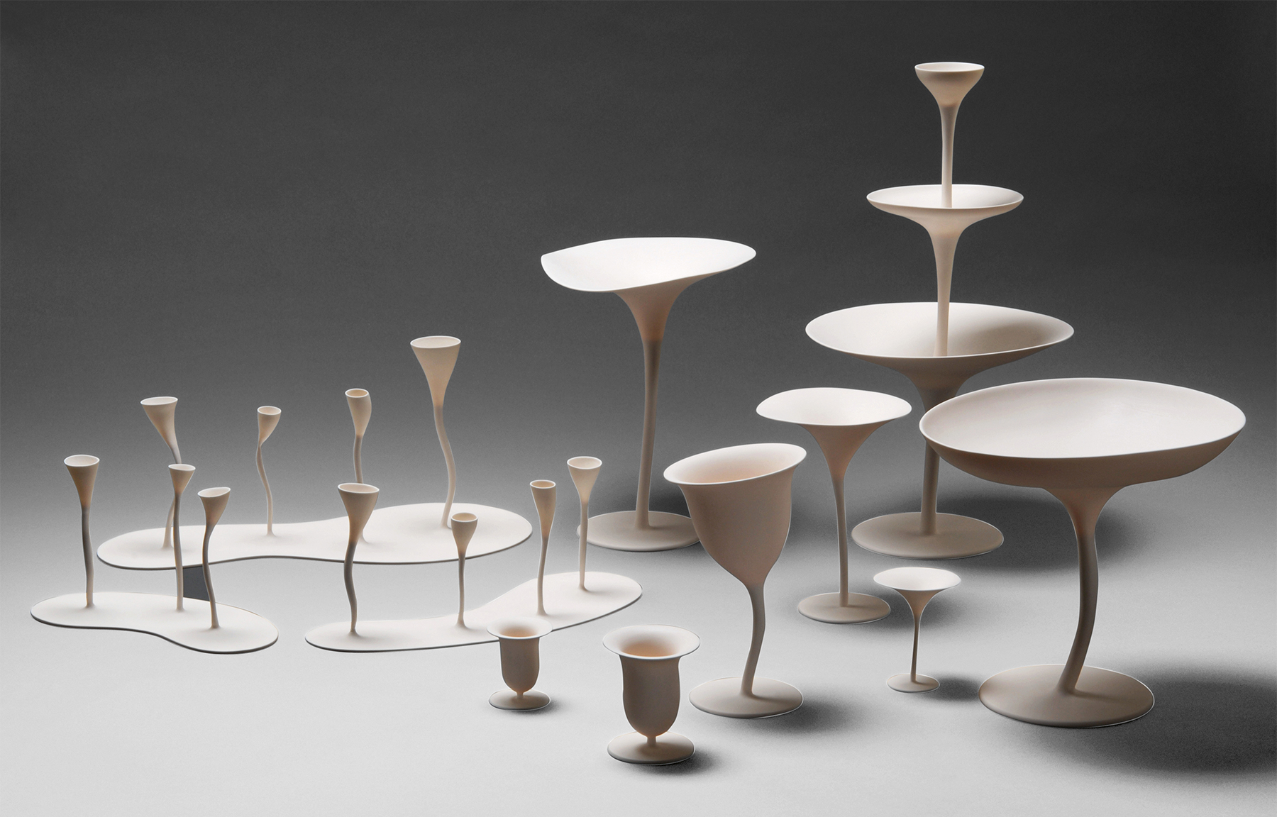 A collection of abstract white sculptures of varying heights and shapes stands against a gray background. These sculptures resemble elongated wine glasses, bowls, and vases with thin, wavy stems and wide, fluid rims, giving a surreal and organic appearance.