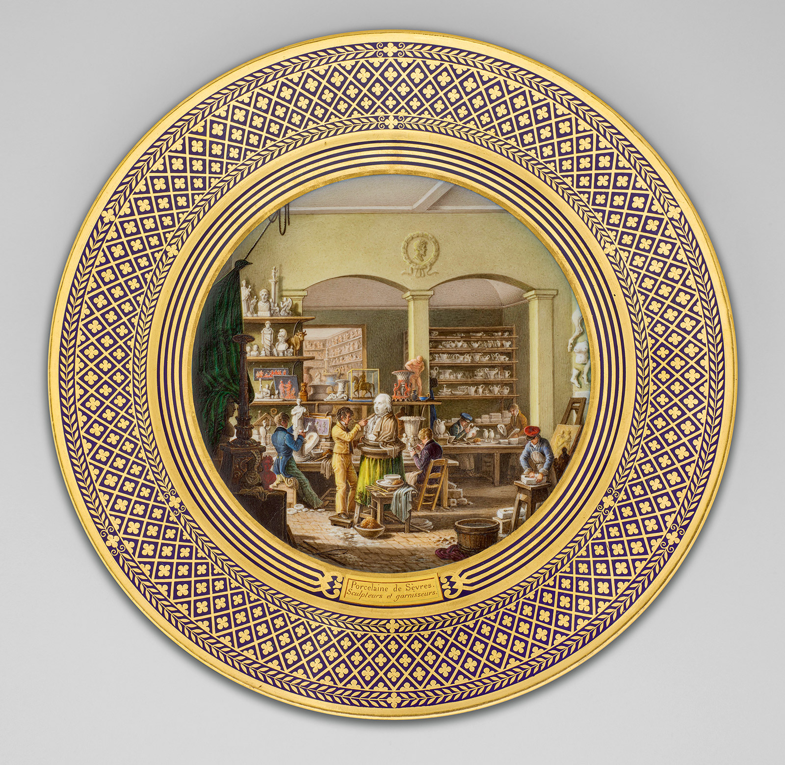 An ornate decorative plate featuring a detailed scene of an 18th-century workshop. The scene portrays artisans crafting pottery and engaging in various tasks. The plate's edge has an intricate gold and purple geometric pattern.