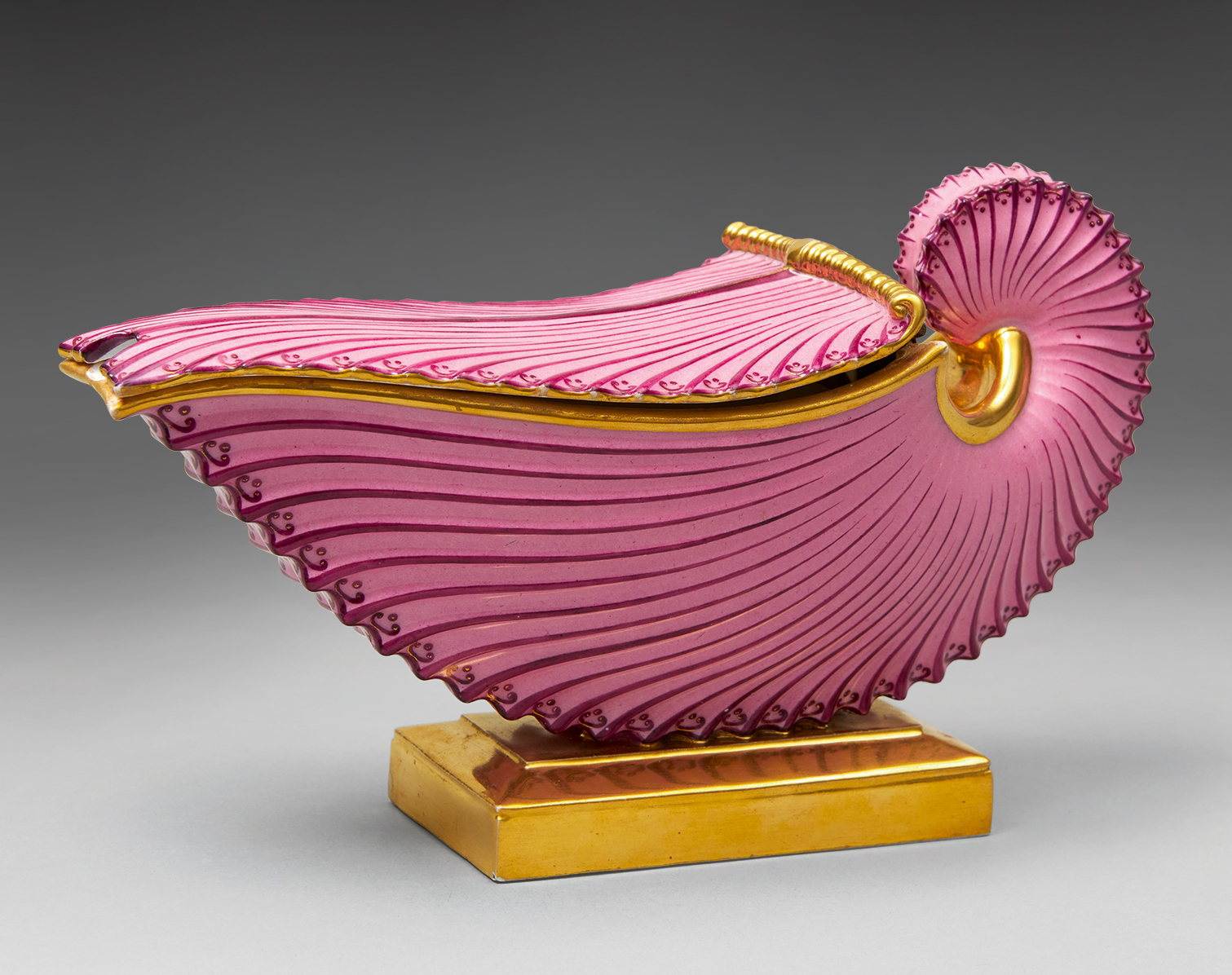 A decorative object shaped like a nautilus shell, featuring a vivid purple exterior with gold accents and elaborate ridged detailing. It rests on a rectangular golden base against a neutral background.