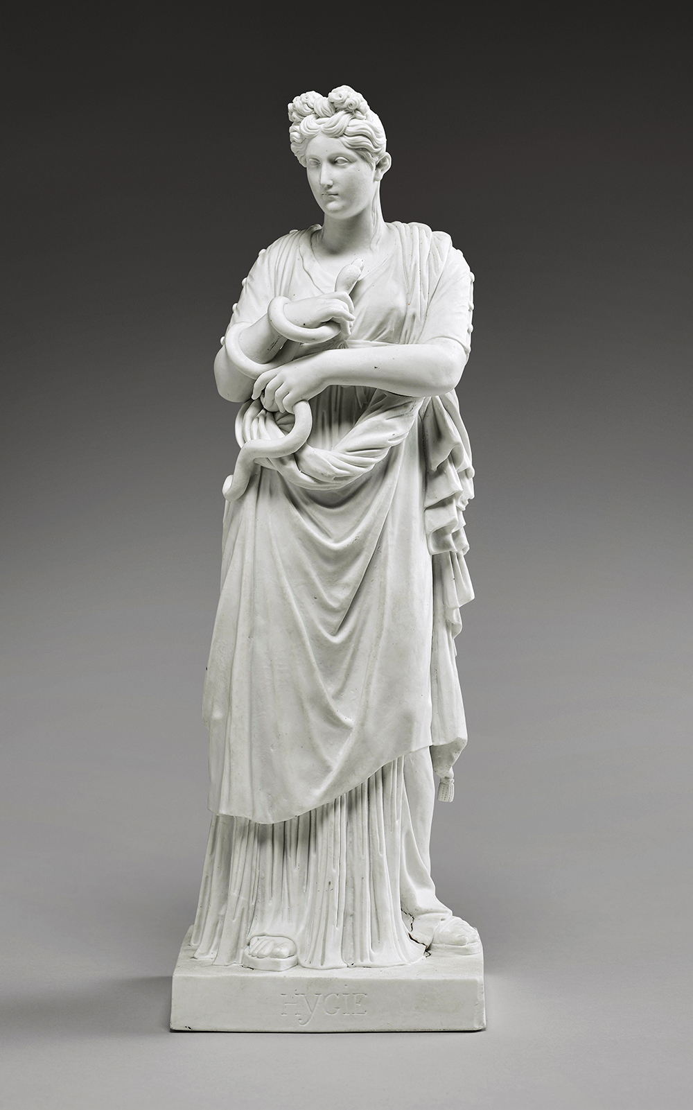 A marble statue depicts a woman standing in a flowing gown, holding a coiled snake in her left arm. She has an ornate hairstyle and her gaze is directed solemnly forward. The sculpture rests on a rectangular base inscribed with Greek characters.