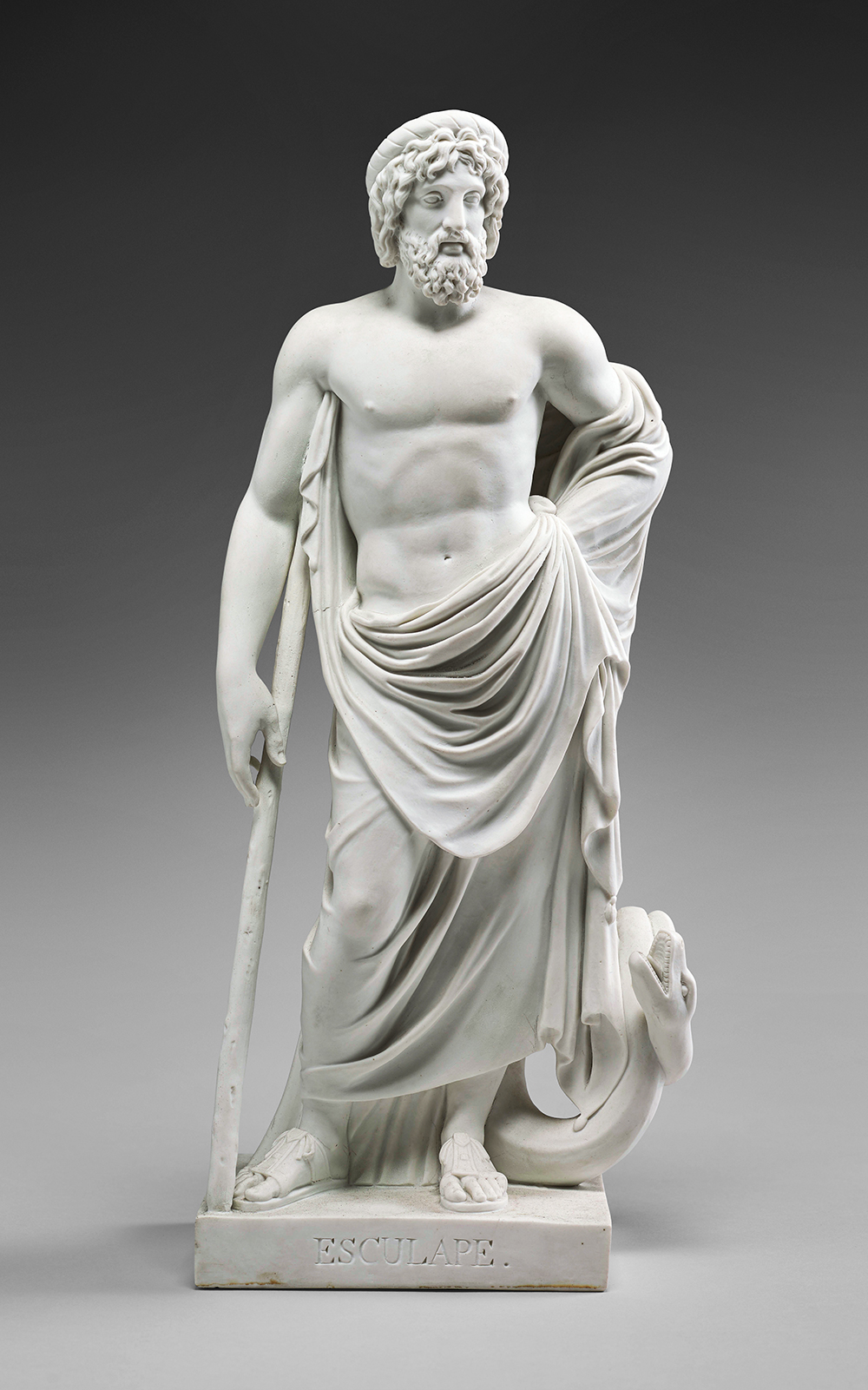 A white marble statue of a bearded, muscular man draped in a cloth. He stands with a staff in his right hand, alongside a snake coiled at his feet. The base of the statue is inscribed with "ESCULAPE." The background is plain and gray.