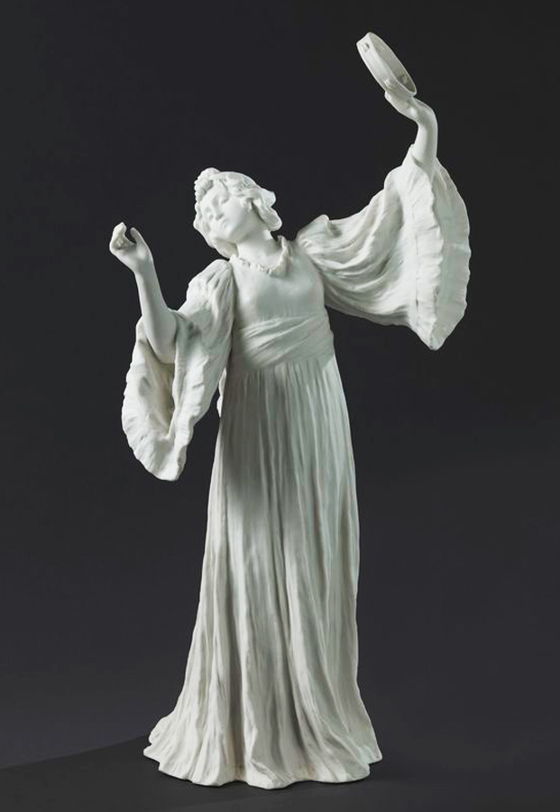 A white ceramic sculpture of a woman in a flowing gown, dancing with arms raised. She holds a small tambourine in one hand. The figure has detailed, ruffled sleeves and a serene expression. The background is dark, highlighting the statue.