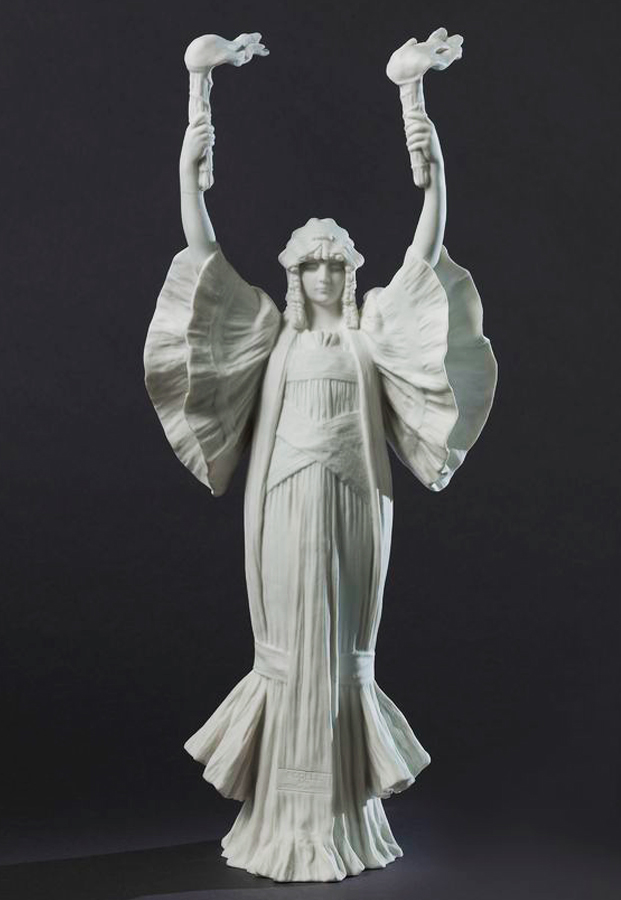 A white ceramic sculpture of a woman in a flowing dress with large sleeves, arms raised, holding two birds. The figure has intricate detailing and stands against a dark background.