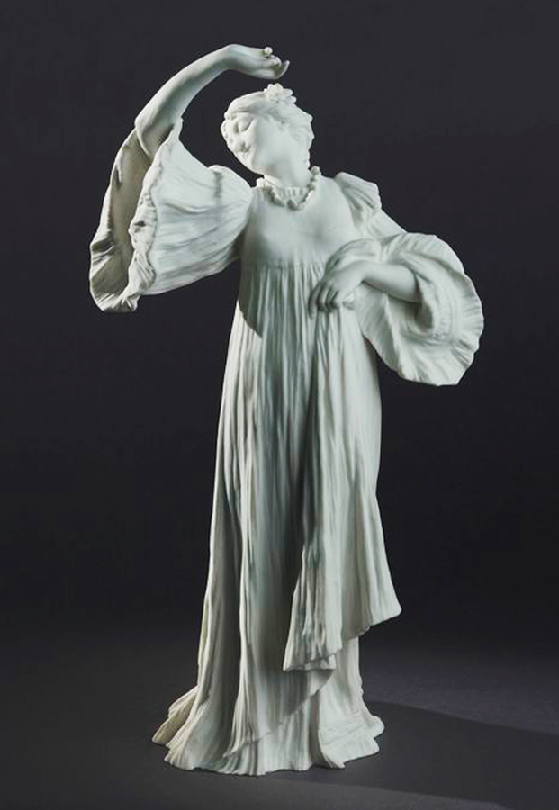 A gracefully posed statue depicts a woman in a flowing gown with voluminous sleeves. Her right arm is raised, and her head tilts back, conveying a sense of elegance and movement. The sculpture is set against a dark background.