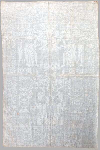 A white linen napkin with a symmetrical pattern featuring a floral border surrounding two distinct scenes. The top depicts a man on a stool conversing with a winged angel while a woman stands in the background. The second is of a man on his knees holding an incense burner. A ram is visible in a thicket above him.