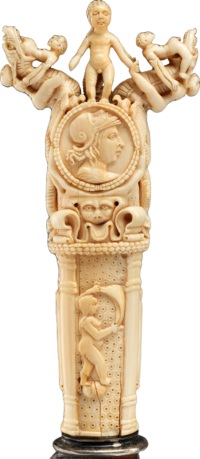 Detail of a carving knife's ivory handle featuring human and animal figures, a face in profile wearing a helmet, and a nude woman holding a billowing piece of fabric.