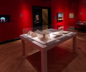 Detail of the Folding by the Book section of the "Staging the Table in Europe 1500-1800" exhibition installation featuring a case of folded napkin examples. In the background several paintings and video screen adorn the room's red walls.