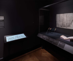 Detail of the Tools of the Trade section of the "Staging the Table in Europe 1500-1800" exhibition installation case featuring various carving tools as well as illustrations and open books with similar tools. An interactive screen displays an image of a table setting with various food items.