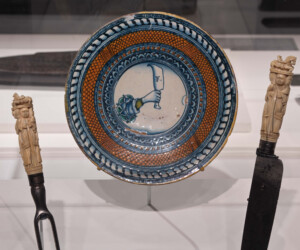 Detail from the "Staging the Table in Europe 1500-1800" exhibition showing a plate with orange and blue concentric designs. At the center is an illustration of a hand holding a cleaver with the letter "f". A metal fork and knife with carved ivory hilts are displayed on either side of the plate.