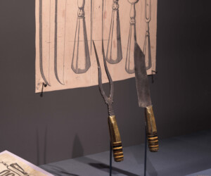 Detail from the "Staging the Table in Europe 1500-1800" exhibition featuring a carving knife and fork with wooden handles in front of a print of other period carving tools.