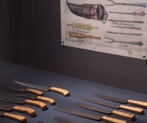 Detail from the "Staging the Table in Europe 1500-1800" exhibition featuring a collection of carving knives with wooden handles. Above the knives is a reproduction of a drawing of a carving tool carying case.