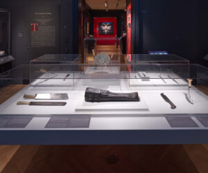 Detail of the Carving by the Book section of the "Staging the Table in Europe 1500-1800" exhibition, featuring multiple display cases with carving tools and an ornate carrying case. Seen in the distance is another room of the exhibition and a double-headed eagle made out of folded linens set against a red wall.