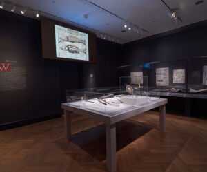 Detail of The Carver section of the "Staging the Table in Europe 1500-1800" exhibition featuring various carving tools enclosed in cases. In the background, open books and illustrations are presented in a case and a large screen projection displays an illustration of a method for carving a fish.