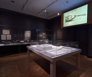 Detail from the "Staging the Table in Europe 1500-1800" exhibition featuring multiple display cases with carving tools. In the background, open books and illustrations are presented in a long case and a large screen projection displays an illustration of a method for carving a boar.