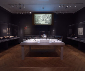 Detail from the "Staging the Table in Europe 1500-1800" exhibition featuring multiple display cases with carving tools. In the background, open books and illustrations are presented in a long cases and a large screen projection displays an illustration of a dining scene in a garden.