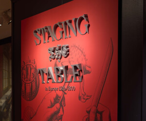 Picture of the title wall for the exhibition "Staging the Table in Europe 1500-1800." The 3D letters are set agains a a red background featuring an illustration of two hands carving a bird.