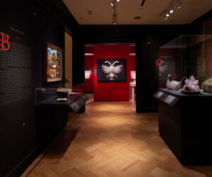 Detail of the "Staging the Table in Europe 1500-1800" exhibition installation featuring several objects in cases. In the center is a large framed double-headed eagle constructed from linen against a red wall.
