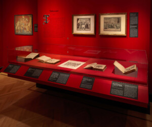 Detail of the Tablescapes section of the "Staging the Table in Europe 1500-1800" exhibition installation featuring several prints and a case of open books on a red background.