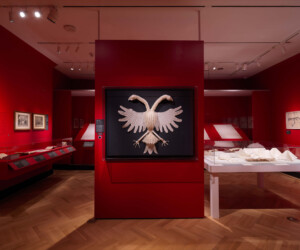 Detail of the Folding by the Book section of the "Staging the Table in Europe 1500-1800" exhibition installation featuring a large double-headed eagle made out of linen on a dark background set on a red wall. The eagle display is flanked by various other objects in cases.