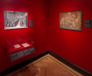 Detail of the Tablescapes section of the "Staging the Table in Europe 1500-1800" exhibition installation. On the left, a glass case contains an open book and manuscript. Two large framed pictures depict period dinning hall scenes.