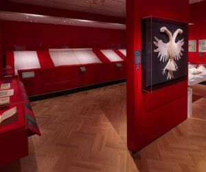 Detail of the Folding by the Book section of the "Staging the Table in Europe 1500-1800" exhibition installation featuring a large double-headed eagle made out of linen on a dark background set on a red wall. The eagle display is flanked by various other objects in cases.